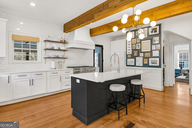 kitchen with white cabinetry, beamed ceiling, premium range hood, pendant lighting, and a center island with sink