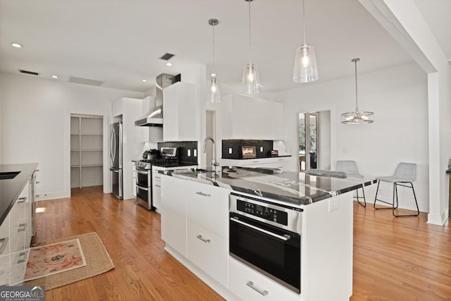 kitchen with white cabinets, appliances with stainless steel finishes, decorative light fixtures, and an island with sink