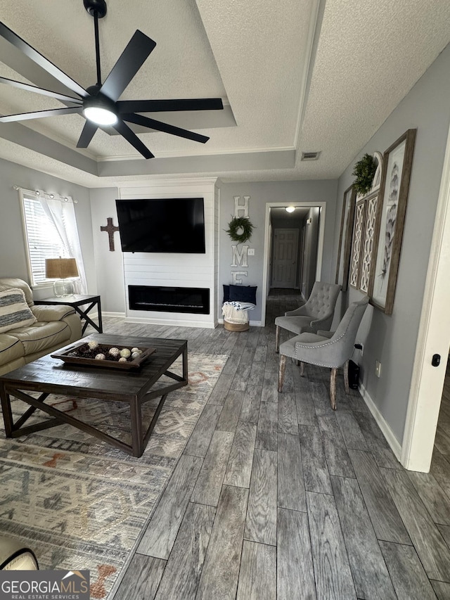 living room with a textured ceiling, ceiling fan, a tray ceiling, and a large fireplace