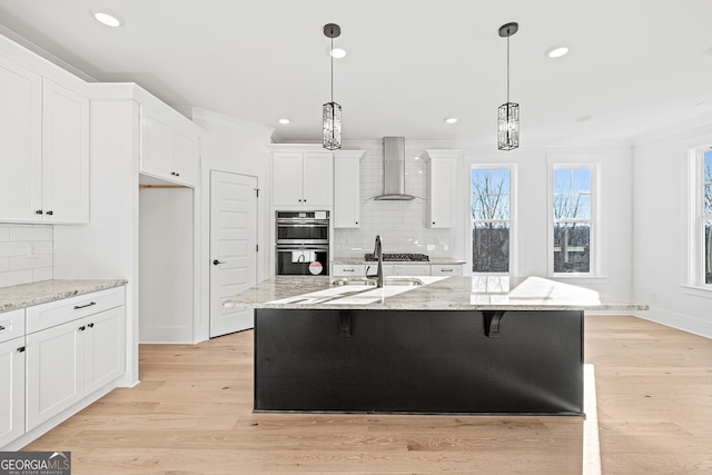 kitchen featuring decorative backsplash, stainless steel appliances, a kitchen island with sink, wall chimney range hood, and light hardwood / wood-style flooring