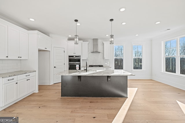 kitchen featuring a center island with sink, light hardwood / wood-style flooring, wall chimney exhaust hood, decorative light fixtures, and white cabinetry