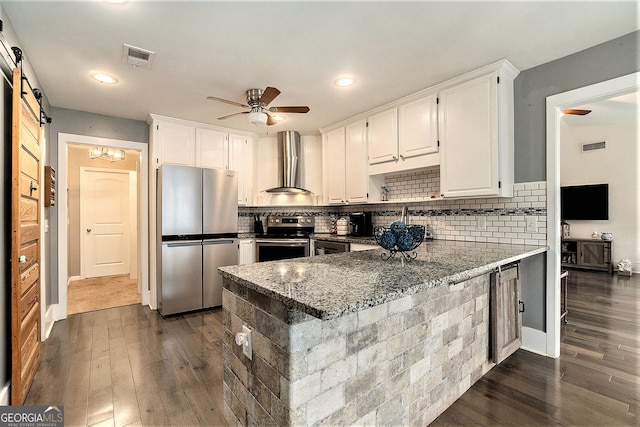 kitchen with stainless steel appliances, wall chimney range hood, a barn door, white cabinets, and dark stone counters