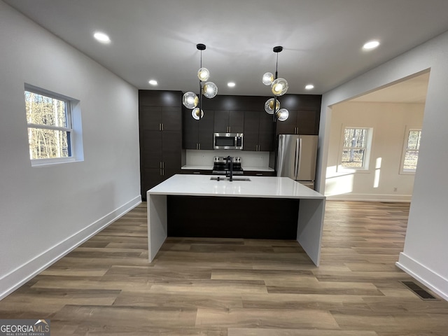 kitchen featuring appliances with stainless steel finishes, decorative light fixtures, sink, light hardwood / wood-style floors, and a center island with sink