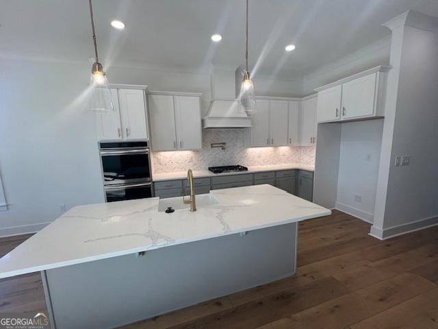 kitchen with pendant lighting, white cabinets, a center island with sink, light stone counters, and custom range hood