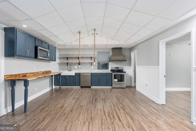 kitchen with wall chimney range hood, sink, blue cabinets, light hardwood / wood-style floors, and stainless steel appliances