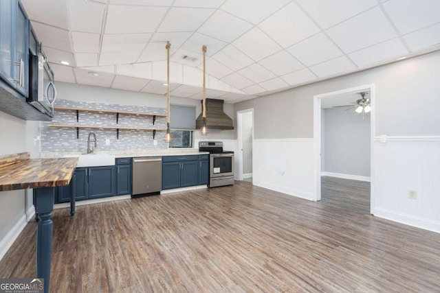 kitchen with wall chimney exhaust hood, dark hardwood / wood-style flooring, blue cabinets, pendant lighting, and appliances with stainless steel finishes