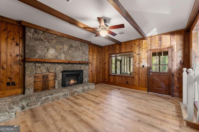 unfurnished living room featuring wood walls, a stone fireplace, ceiling fan, light wood-type flooring, and beam ceiling