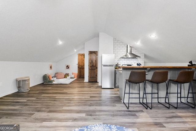 interior space featuring stainless steel refrigerator, wall chimney exhaust hood, a kitchen breakfast bar, light hardwood / wood-style floors, and vaulted ceiling