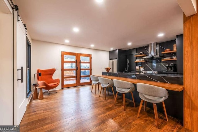 kitchen with kitchen peninsula, dark hardwood / wood-style flooring, high end fridge, wall chimney range hood, and a barn door