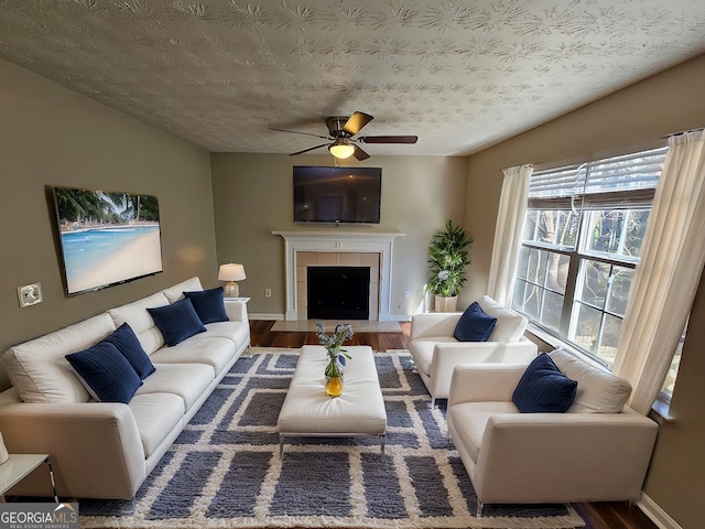 living room with a textured ceiling, ceiling fan, a tiled fireplace, and wood-type flooring