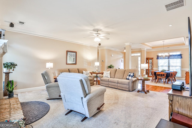 living room with ceiling fan with notable chandelier, a raised ceiling, crown molding, light colored carpet, and decorative columns