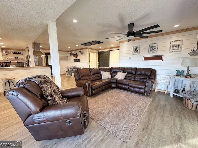 living room featuring wooden walls, ceiling fan, a barn door, a textured ceiling, and light hardwood / wood-style floors