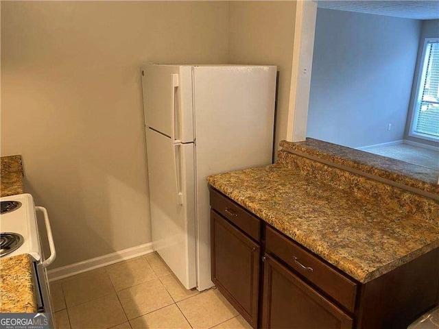 kitchen featuring dark brown cabinetry, white fridge, stone countertops, light tile patterned floors, and range