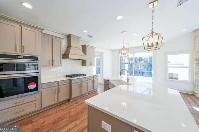 kitchen with a kitchen island with sink, sink, hanging light fixtures, custom range hood, and stainless steel appliances