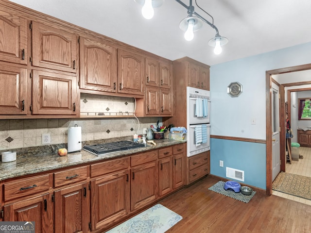 kitchen featuring light wood-type flooring, tasteful backsplash, dark stone counters, stainless steel gas cooktop, and double oven