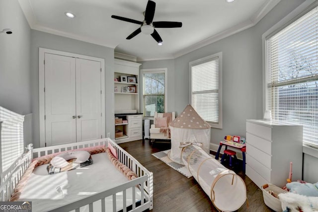 bedroom with ornamental molding, ceiling fan, dark wood-type flooring, a crib, and a closet