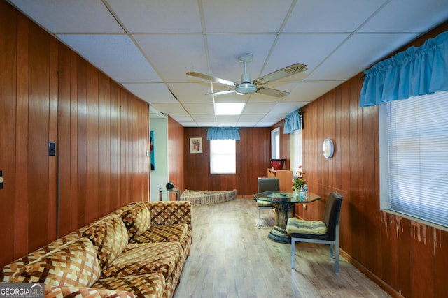 living room with ceiling fan, a paneled ceiling, wooden walls, and light hardwood / wood-style flooring