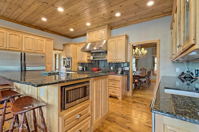 kitchen with a center island with sink, stainless steel microwave, dark stone countertops, and light brown cabinetry