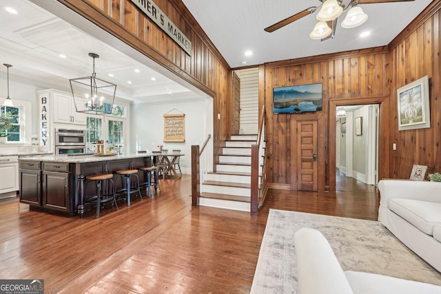 living room with a raised ceiling, dark hardwood / wood-style floors, wooden walls, ceiling fan with notable chandelier, and ornamental molding