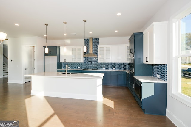 kitchen featuring wall chimney range hood, tasteful backsplash, an island with sink, pendant lighting, and white cabinets