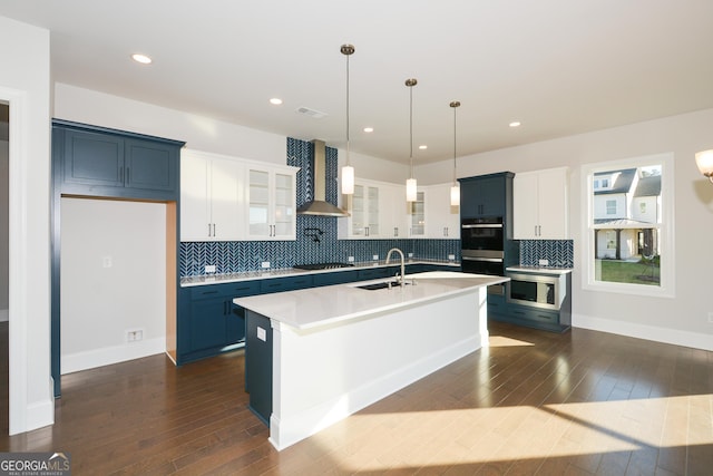 kitchen with sink, wall chimney exhaust hood, an island with sink, decorative light fixtures, and white cabinetry