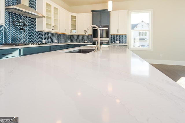 kitchen with wall chimney range hood, sink, light stone counters, white cabinetry, and stainless steel appliances