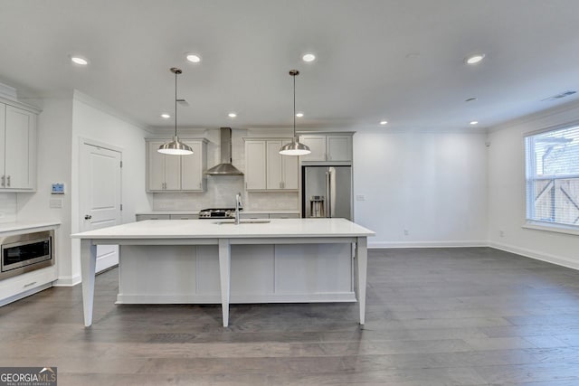 kitchen with dark wood-type flooring, stainless steel appliances, wall chimney range hood, pendant lighting, and a center island with sink