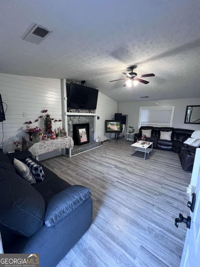 living room with a stone fireplace, wooden walls, ceiling fan, a textured ceiling, and wood-type flooring