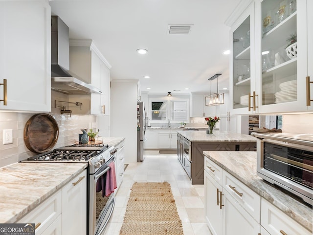 kitchen with appliances with stainless steel finishes, light stone counters, wall chimney range hood, white cabinetry, and pendant lighting