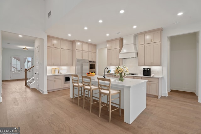 kitchen with a breakfast bar area, light hardwood / wood-style flooring, appliances with stainless steel finishes, an island with sink, and custom range hood