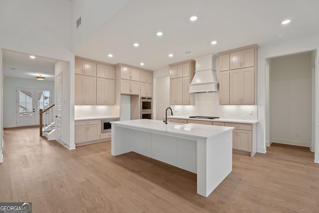 kitchen with sink, stainless steel appliances, custom range hood, an island with sink, and light wood-type flooring