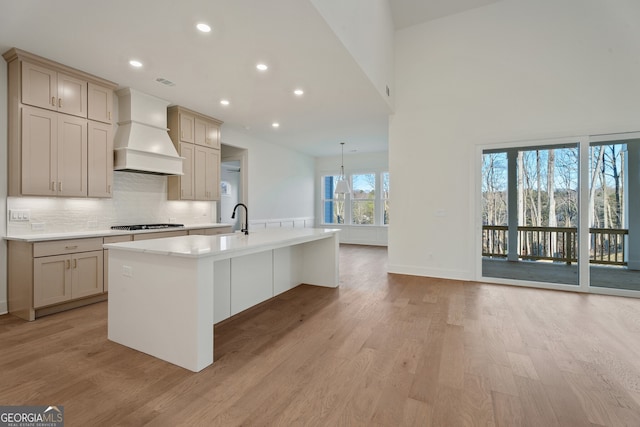 kitchen featuring stainless steel gas cooktop, custom exhaust hood, a center island with sink, light hardwood / wood-style floors, and backsplash