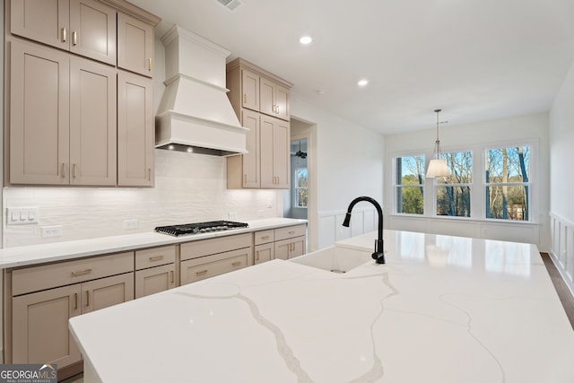 kitchen featuring stainless steel gas cooktop, sink, custom exhaust hood, hanging light fixtures, and light stone countertops