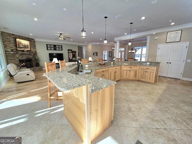 kitchen with light brown cabinets, ceiling fan with notable chandelier, a kitchen breakfast bar, sink, and a brick fireplace