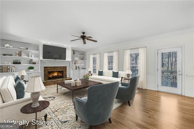 living room featuring a stone fireplace, crown molding, ceiling fan, built in features, and wood-type flooring