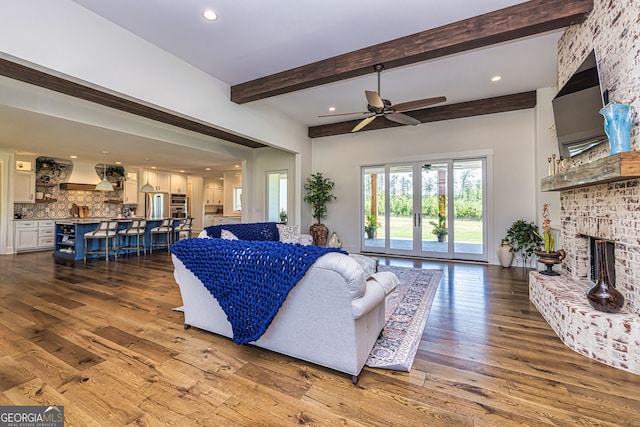 living room featuring beam ceiling, ceiling fan, wood-type flooring, and a brick fireplace