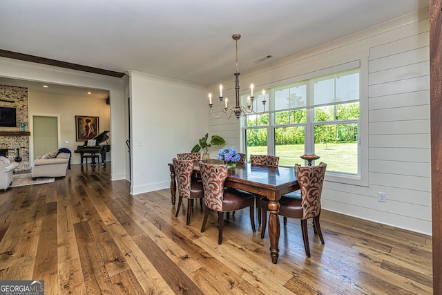 dining room featuring an inviting chandelier, wooden walls, hardwood / wood-style flooring, ornamental molding, and a large fireplace