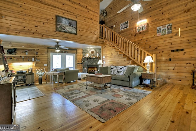 living room featuring wood walls, a stone fireplace, ceiling fan, a towering ceiling, and light hardwood / wood-style floors