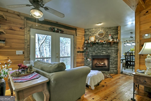 living room featuring french doors, light wood-type flooring, ceiling fan, wooden walls, and a stone fireplace