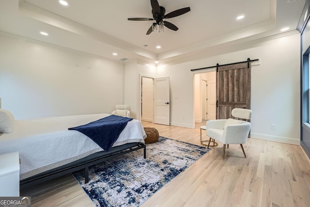 bedroom featuring ceiling fan, a barn door, light wood-type flooring, and a tray ceiling