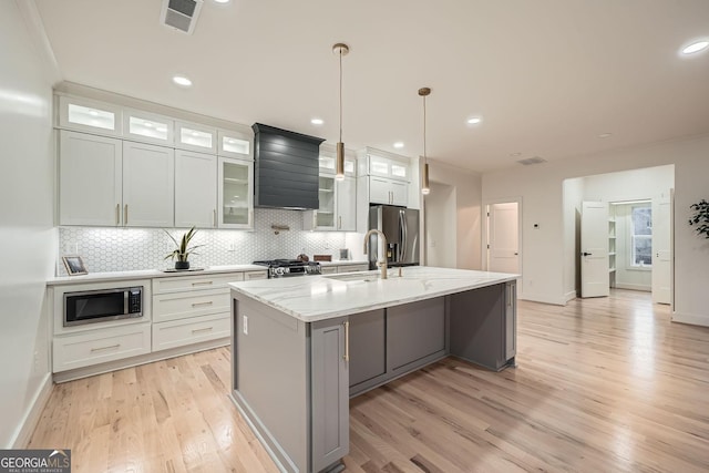 kitchen with pendant lighting, appliances with stainless steel finishes, white cabinetry, a center island with sink, and wall chimney exhaust hood