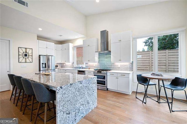 kitchen with light stone countertops, appliances with stainless steel finishes, wall chimney exhaust hood, white cabinetry, and a kitchen island