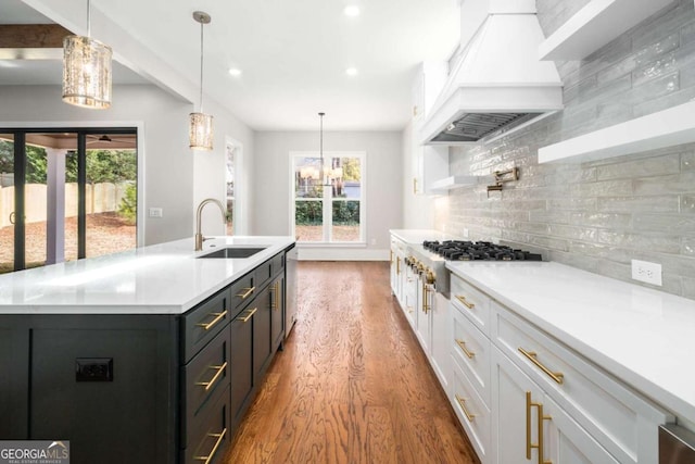 kitchen featuring premium range hood, decorative light fixtures, sink, white cabinets, and a center island with sink