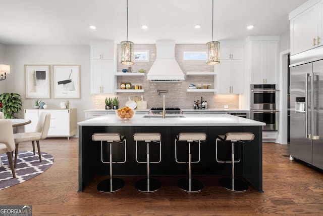 kitchen featuring appliances with stainless steel finishes, custom range hood, hanging light fixtures, and a center island with sink