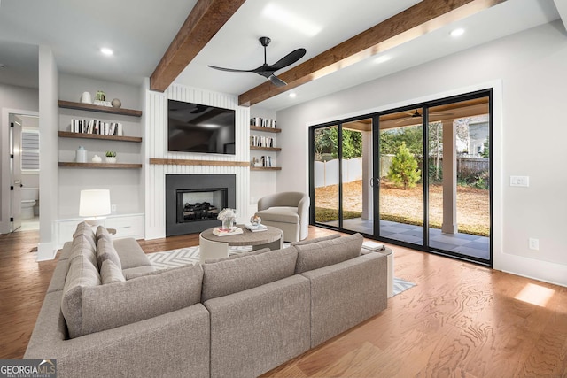 living room with built in features, ceiling fan, beam ceiling, a large fireplace, and light wood-type flooring