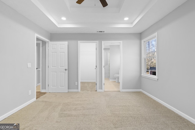 unfurnished bedroom featuring connected bathroom, ceiling fan, a tray ceiling, a walk in closet, and light carpet