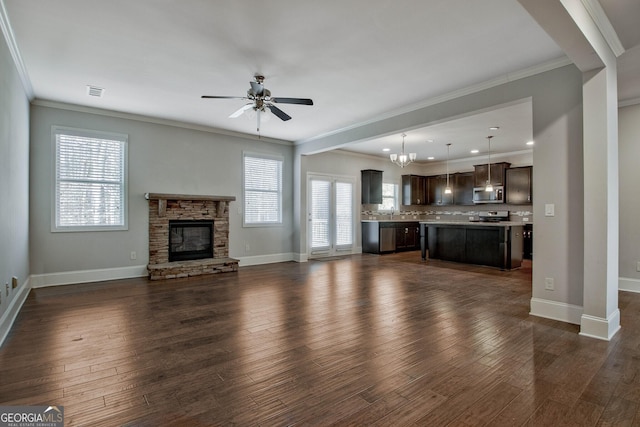 unfurnished living room with ceiling fan with notable chandelier, dark wood-type flooring, a stone fireplace, and crown molding