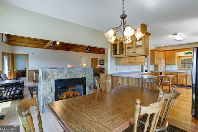 dining room with ceiling fan with notable chandelier, beam ceiling, light wood-type flooring, and a stone fireplace