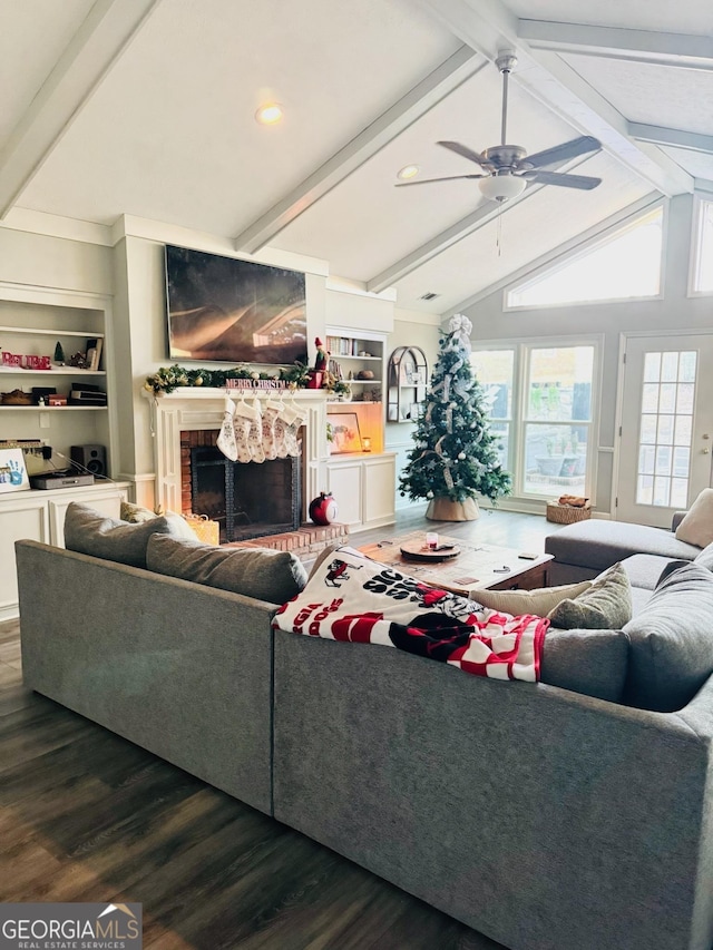 living room featuring lofted ceiling with beams, ceiling fan, dark wood-type flooring, a brick fireplace, and built in shelves