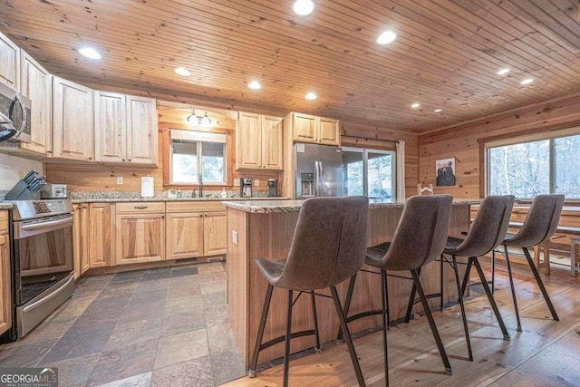 kitchen with wood walls, light stone countertops, light brown cabinetry, appliances with stainless steel finishes, and a kitchen island